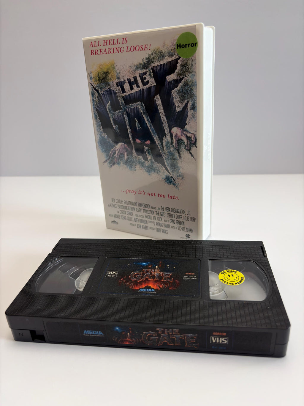 May include: A white VHS case for the movie The Gate with the text All Hell Is Breaking Loose and a graphic of a dark pit, alongside a black VHS tape with a label featuring the title The Gate and a yellow Be Kind Rewind smiley face sticker.