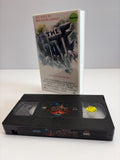 May include: A white VHS case for the movie The Gate with the text All Hell Is Breaking Loose and a graphic of a dark pit, alongside a black VHS tape with a label featuring the title The Gate and a yellow Be Kind Rewind smiley face sticker.